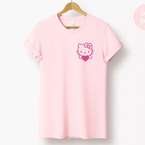 May include: A light pink t-shirt with a graphic of a cartoon cat holding a heart. The cat has a bow on its head. The t-shirt is hanging on a wooden hanger.
