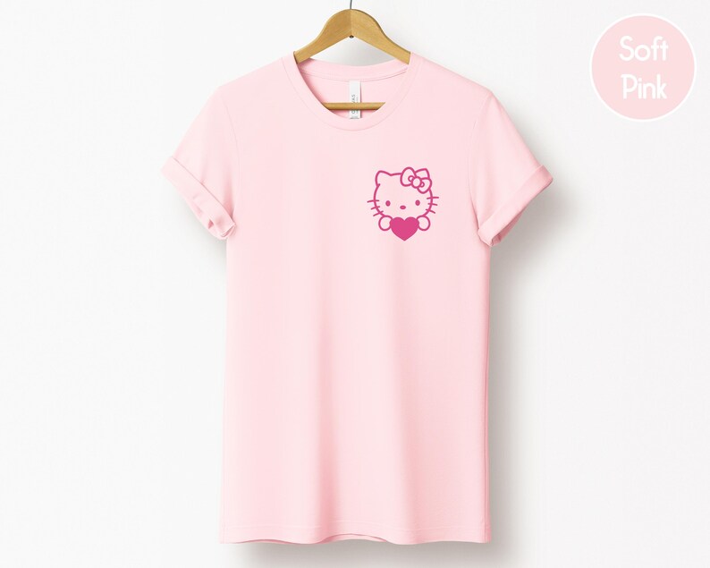 May include: A light pink t-shirt with a graphic of a cartoon cat holding a heart. The cat has a bow on its head. The t-shirt is hanging on a wooden hanger.
