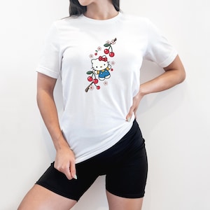 Op de afbeelding: Wit T-shirt met korte mouwen met een cartoonkarakterillustratie. Het ontwerp omvat een rode strik, blauwe overalls en een kersenboomtak met rode kersen en witte en roze bloemen.