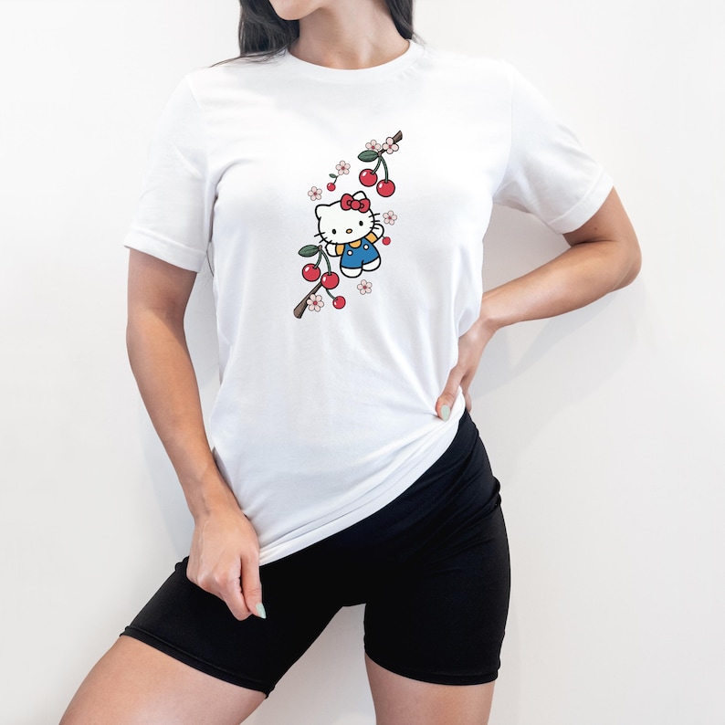 Op de afbeelding: Wit T-shirt met korte mouwen met een cartoonkarakterillustratie. Het ontwerp omvat een rode strik, blauwe overalls en een kersenboomtak met rode kersen en witte en roze bloemen.