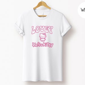Op de afbeelding: Wit T-shirt met een roze opdruk met het woord "LOVEY", een Hello Kitty-personage en het woord "Hellokitty". Het T-shirt hangt aan een houten hanger tegen een witte achtergrond.
