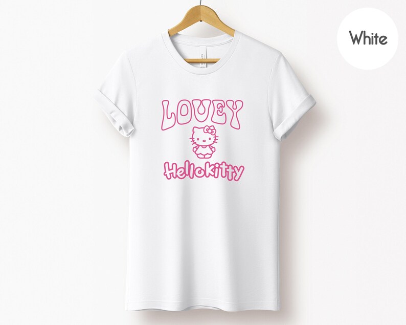 Op de afbeelding: Wit T-shirt met een roze opdruk met het woord "LOVEY", een Hello Kitty-personage en het woord "Hellokitty". Het T-shirt hangt aan een houten hanger tegen een witte achtergrond.