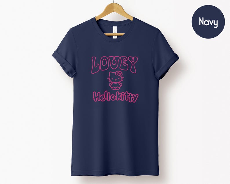 Op de afbeelding: Marineblauw T-shirt met opgerolde mouwen. Het shirt heeft de tekst "LOUEY" boven een Hello Kitty-afbeelding, met "Hellokitty" eronder in roze. Het T-shirt hangt aan een houten hanger tegen een witte achtergrond.