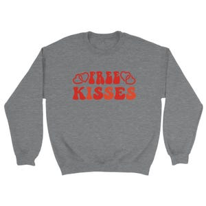 May include: A grey crew neck sweatshirt featuring the words "FREE KISSES" in a retro red and orange font. The word "FREE" is flanked by heart shapes. A casual, comfortable garment.