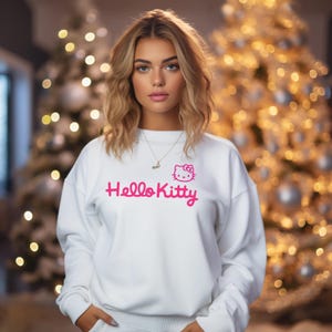 May include: A white sweatshirt featuring the pink text "Hello Kitty" and a matching pink Hello Kitty graphic. The sweatshirt is part of a set. The background is blurred with Christmas lights.