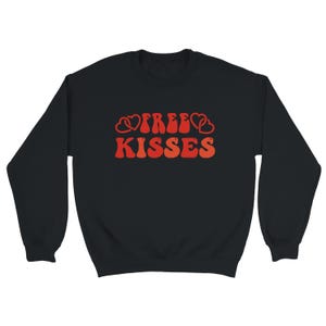May include: Black sweatshirt featuring the text "FREE KISSES" in a retro style. The lettering is in shades of red and orange, with heart motifs. A relaxed, comfortable garment.