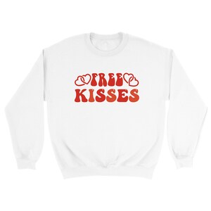 May include: White sweatshirt featuring the text "FREE KISSES" in red, retro font, with heart motifs. A relaxed-fit top, suitable for Valentine's Day or general wear. A comfortable, casual garment.