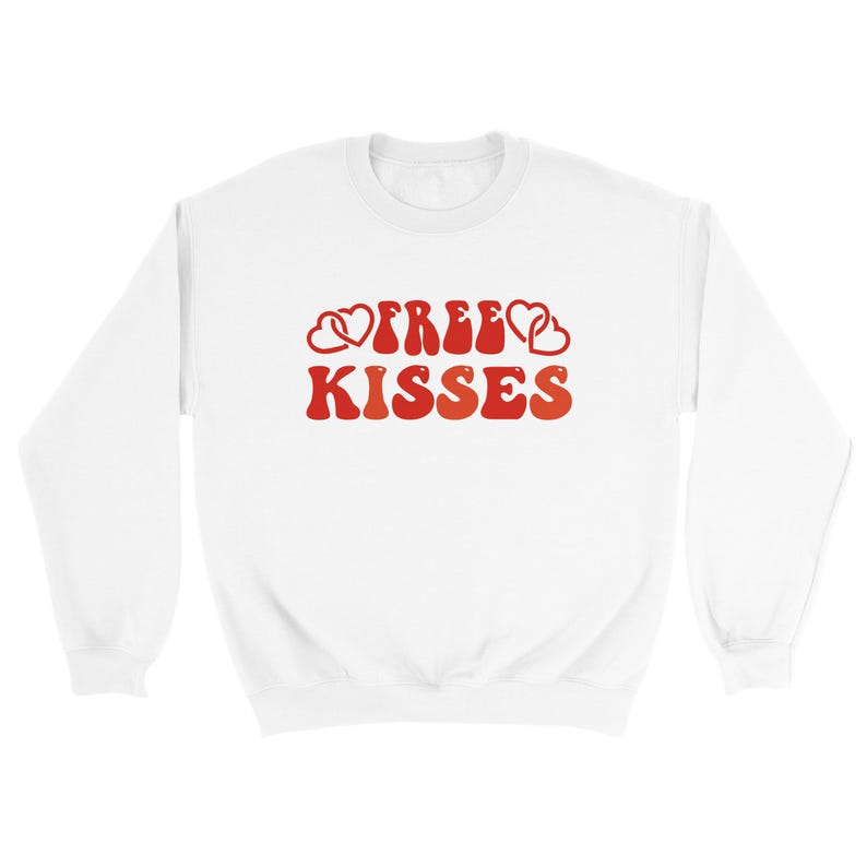 May include: White sweatshirt featuring the text "FREE KISSES" in red, retro font, with heart motifs. A relaxed-fit top, suitable for Valentine's Day or general wear. A comfortable, casual garment.