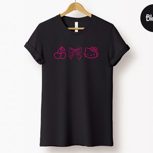 Pu&ograve; includere: T-shirt nera con un design rosa a forma di ciliegia, fiocco e contorno di Hello Kitty. L'indumento &egrave; appeso a una gruccia di legno su sfondo bianco. La parola "Black" &egrave; in un cerchio nero.