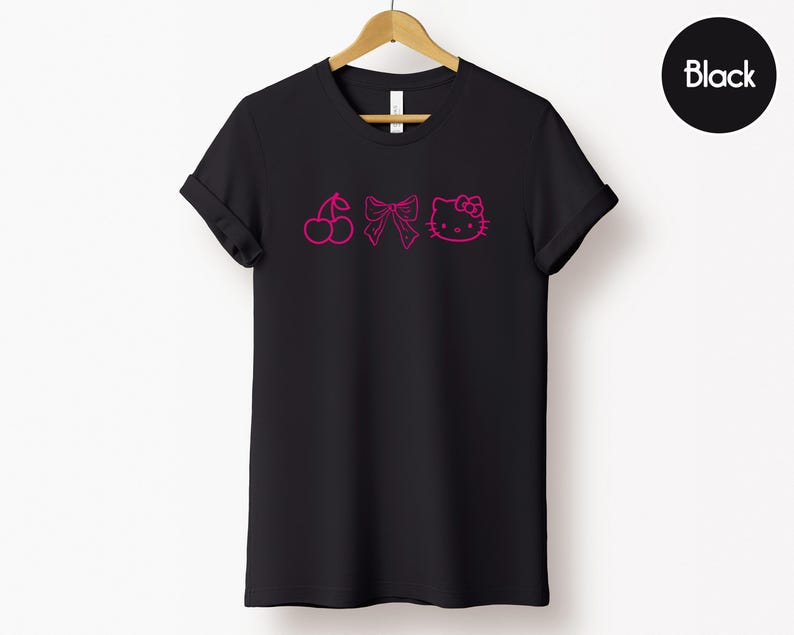 Pu&ograve; includere: T-shirt nera con un design rosa a forma di ciliegia, fiocco e contorno di Hello Kitty. L'indumento &egrave; appeso a una gruccia di legno su sfondo bianco. La parola "Black" &egrave; in un cerchio nero.
