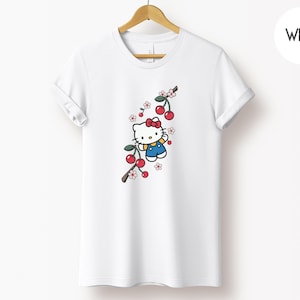Op de afbeelding: Wit T-shirt met een cartoonfiguur met een rode strik en blauwe overall, met een kersentakontwerp. De kersen zijn rood en de bloemen roze en wit. Het T-shirt hangt aan een houten hanger.