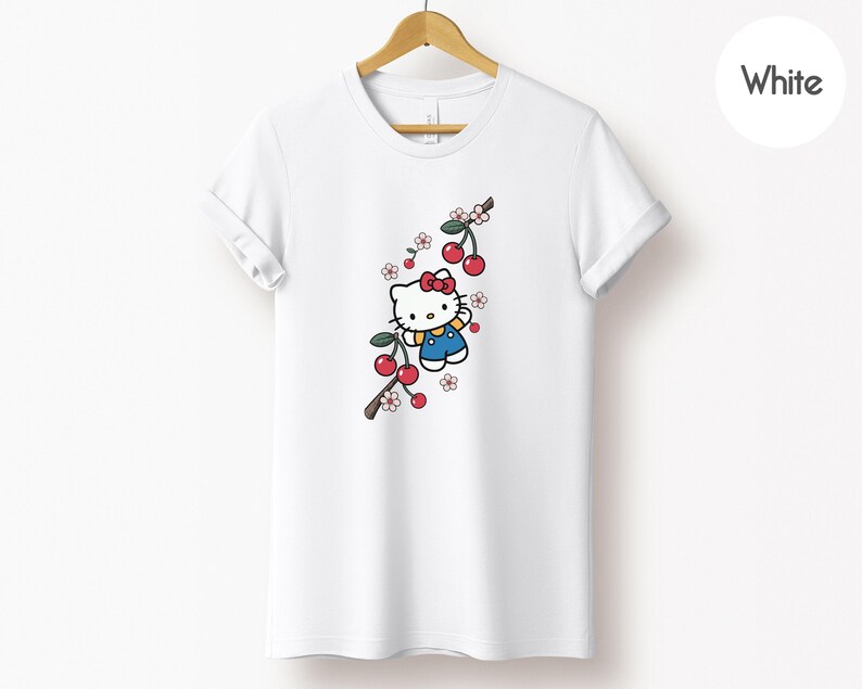 Op de afbeelding: Wit T-shirt met een cartoonfiguur met een rode strik en blauwe overall, met een kersentakontwerp. De kersen zijn rood en de bloemen roze en wit. Het T-shirt hangt aan een houten hanger.