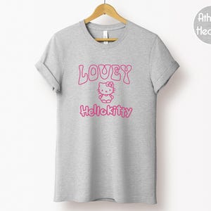 Op de afbeelding: Een heather grijze t-shirt met de roze tekst "LOVEY HelloKitty" en een Hello Kitty-afbeelding. Het t-shirt hangt aan een houten hanger. De stof is athletic heather.