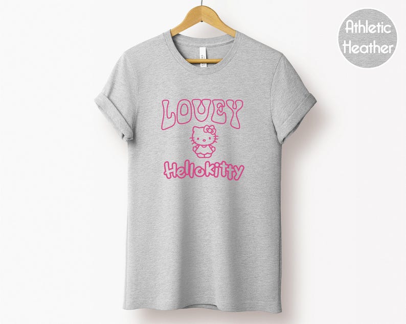 Op de afbeelding: Een heather grijze t-shirt met de roze tekst "LOVEY HelloKitty" en een Hello Kitty-afbeelding. Het t-shirt hangt aan een houten hanger. De stof is athletic heather.