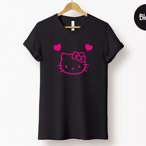 以下が含まれることがあります： ピンクのハローキティのデザインと2つのピンクのハートが付いた黒いTシャツ。「Black」の文字は黒い円の中にあります。Tシャツは木製のハンガーに掛けられています。