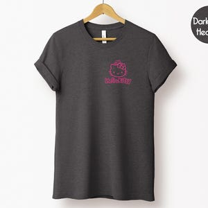 May include: Dark grey heather t-shirt with a pink Hello Kitty graphic on the front. The graphic includes the Hello Kitty character and the text "HelloKitty". The t-shirt is hanging on a wooden hanger.