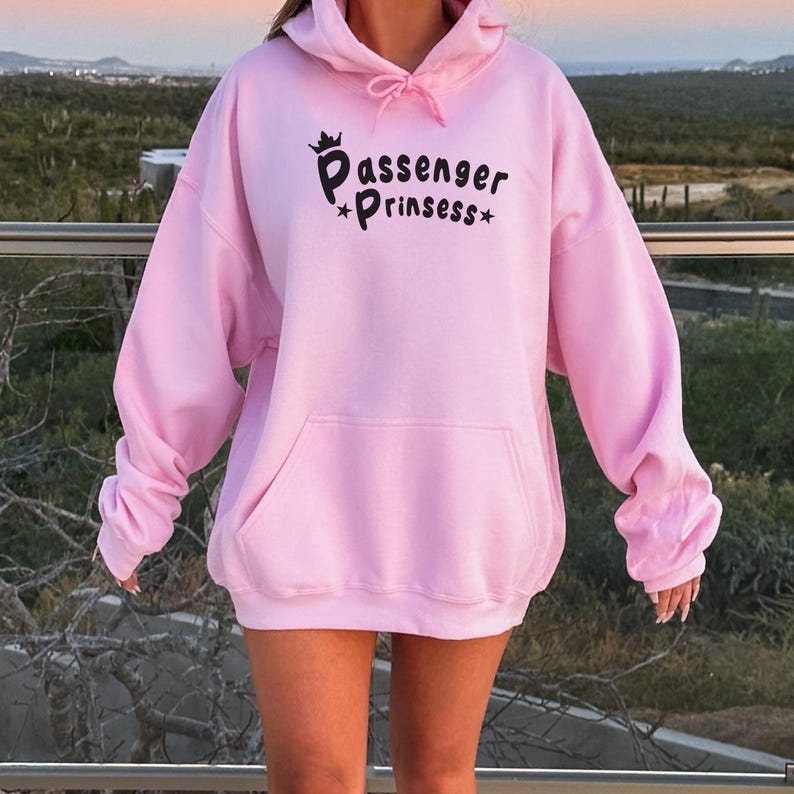 May include: A pale pink hoodie with a front pocket and a drawstring hood. The hoodie displays the text "Passenger Prinssess" in black script with a crown and stars. The sleeves are long and the material looks soft.