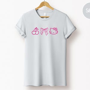 Pu&ograve; includere: T-shirt grigio chiaro con ciliegie, fiocco e Hello Kitty rosa. La maglietta &egrave; appesa a una gruccia di legno su sfondo bianco. Le maniche sono arrotolate.
