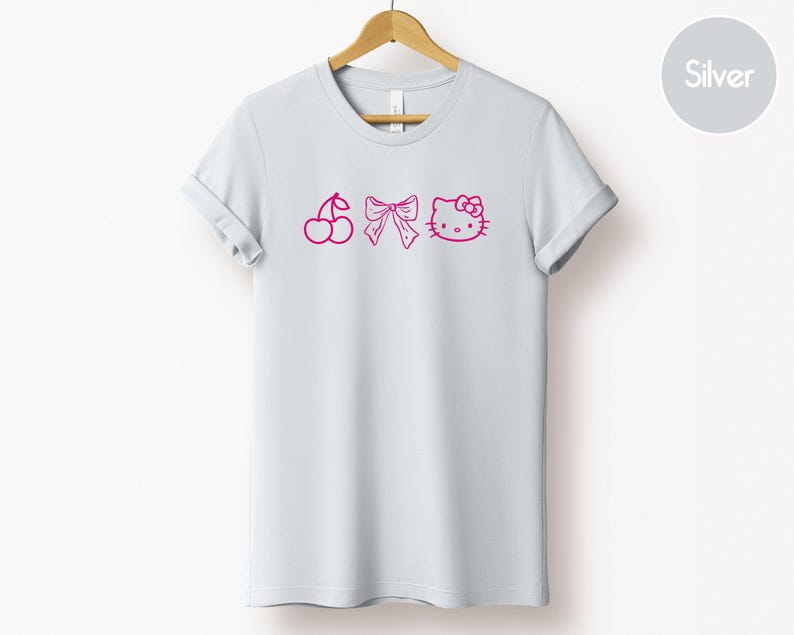 Pu&ograve; includere: T-shirt grigio chiaro con ciliegie, fiocco e Hello Kitty rosa. La maglietta &egrave; appesa a una gruccia di legno su sfondo bianco. Le maniche sono arrotolate.
