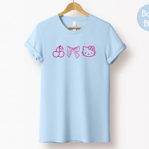 Pu&ograve; includere: T-shirt azzurra con un design rosa a forma di ciliegia, fiocco e volto di gatto. La maglietta &egrave; appesa a una gruccia di legno. Un cerchio azzurro con la scritta "Baby Blue" si trova in alto a destra.