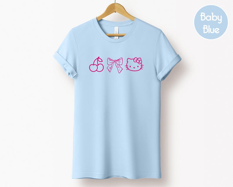 Pu&ograve; includere: T-shirt azzurra con un design rosa a forma di ciliegia, fiocco e volto di gatto. La maglietta &egrave; appesa a una gruccia di legno. Un cerchio azzurro con la scritta "Baby Blue" si trova in alto a destra.