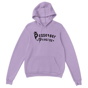 May include: A lilac hoodie with a hood and a front pocket. The hoodie displays the text "Passenger Princess" in black, with a crown above the first word and stars around the second word.