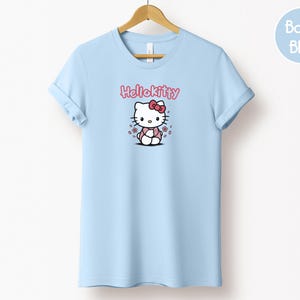 May include: Light blue t-shirt featuring a Hello Kitty graphic. The graphic includes the text "Hello Kitty" in pink above an illustration of the character. The shirt is hanging on a wooden hanger.