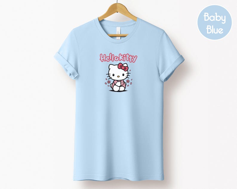May include: Light blue t-shirt featuring a Hello Kitty graphic. The graphic includes the text "Hello Kitty" in pink above an illustration of the character. The shirt is hanging on a wooden hanger.