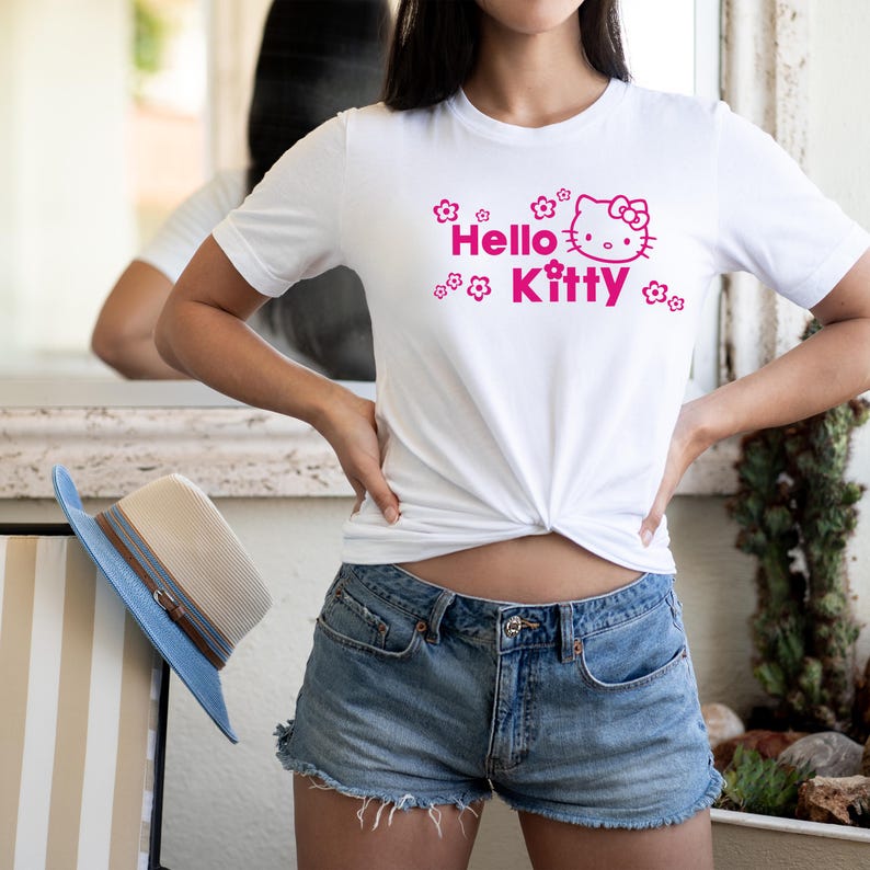 May include: White t-shirt with a pink Hello Kitty design and floral details. The top is knotted at the waist, worn with denim shorts. A blue and beige hat is visible.