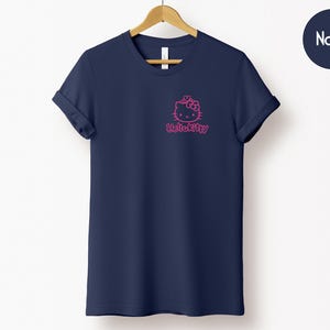 May include: Navy blue t-shirt with a pink Hello Kitty graphic on the front. The t-shirt has short sleeves and a crew neck. The text "Hello Kitty" is below the graphic. The shirt is hanging on a wooden hanger.