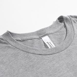 May include: Close-up of a heather gray t-shirt neckline. The shirt has a crew neck and a tag that reads "ANVAS LA CANVAS". The fabric appears soft and the stitching is visible.