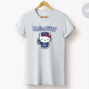 May include: A pale grey t-shirt with a cartoon Hello Kitty character wearing a blue baseball cap and jersey. The text "Hello Kitty" is above the character. The character is holding a blue baseball glove.