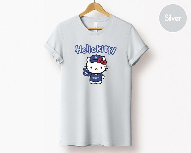May include: A pale grey t-shirt with a cartoon Hello Kitty character wearing a blue baseball cap and jersey. The text "Hello Kitty" is above the character. The character is holding a blue baseball glove.