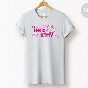 May include: A white t-shirt featuring a pink Hello Kitty graphic and the words "Hello Kitty". The design includes floral elements. The garment is displayed on a wooden hanger.