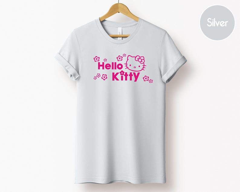 May include: A white t-shirt featuring a pink Hello Kitty graphic and the words "Hello Kitty". The design includes floral elements. The garment is displayed on a wooden hanger.
