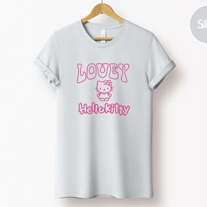 Op de afbeelding: Een lichtgrijs t-shirt met opgerolde mouwen, met de tekst "LOVEY" boven een roze Hello Kitty-afbeelding en het woord "Hellokitty" eronder. Het shirt hangt aan een houten hanger.