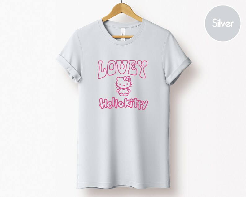 Op de afbeelding: Een lichtgrijs t-shirt met opgerolde mouwen, met de tekst "LOVEY" boven een roze Hello Kitty-afbeelding en het woord "Hellokitty" eronder. Het shirt hangt aan een houten hanger.