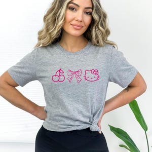 Pu&ograve; includere: Una maglietta grigio melange con una grafica rosa di una ciliegia, un fiocco e Hello Kitty. La maglietta a maniche corte &egrave; realizzata in materiale morbido ed &egrave; annodata in vita. La grafica &egrave; in linea.