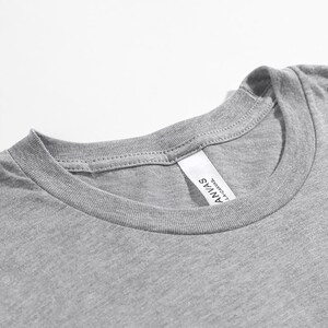 May include: A close-up shot of a heather grey t-shirt neckline. The shirt features a crew neck and a label that reads "ANVAS LA CANVAS". The fabric looks soft and the stitching is visible.