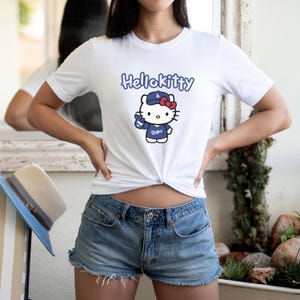 May include: White t-shirt with a Hello Kitty graphic in a Dodgers baseball uniform. The design includes the text "Hello Kitty" in blue script above the character. The character is wearing a blue cap and holding up a peace sign.