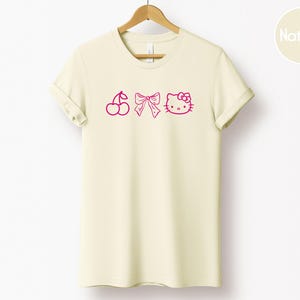 Pu&ograve; includere: T-shirt color crema con ciliegia rosa, fiocco e grafica di Hello Kitty. La maglietta &egrave; appesa a una gruccia di legno. La parola "Natural" &egrave; stampata su un cerchio in alto a destra.