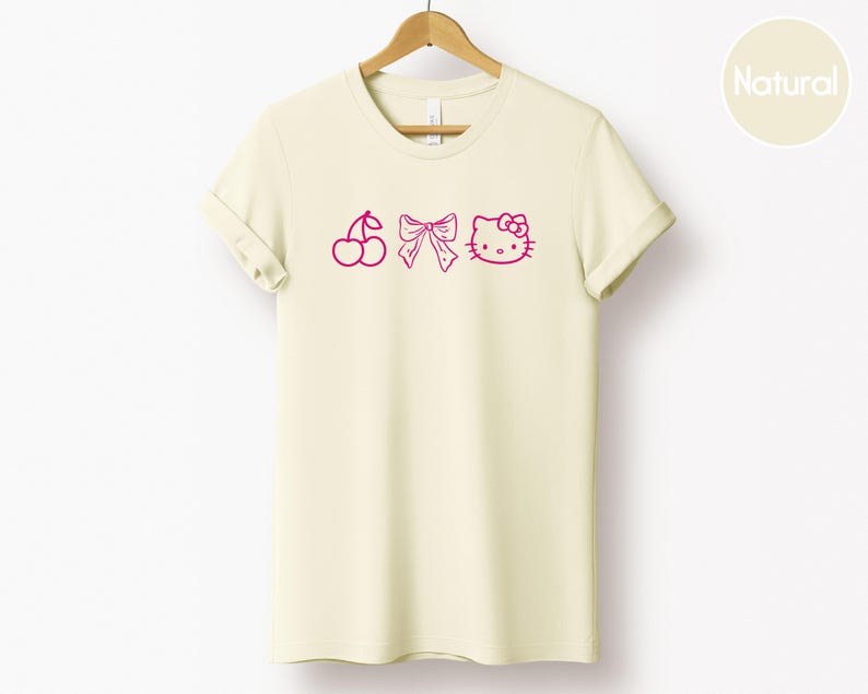 Pu&ograve; includere: T-shirt color crema con ciliegia rosa, fiocco e grafica di Hello Kitty. La maglietta &egrave; appesa a una gruccia di legno. La parola "Natural" &egrave; stampata su un cerchio in alto a destra.