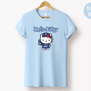 May include: A light blue t-shirt with a Hello Kitty graphic wearing a baseball cap and uniform. The text "Hellokitty" is above the graphic. The shirt is displayed on a wooden hanger.