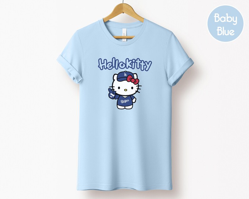 May include: A light blue t-shirt with a Hello Kitty graphic wearing a baseball cap and uniform. The text "Hellokitty" is above the graphic. The shirt is displayed on a wooden hanger.