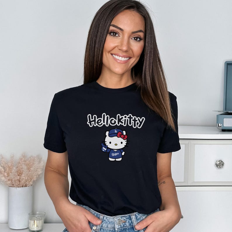May include: Black t-shirt with a Hello Kitty graphic in a baseball uniform. The design includes the text "Hello Kitty" above the character. The character is wearing a baseball cap and uniform.