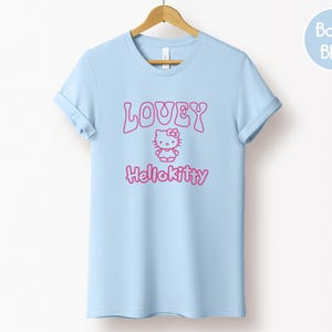 Op de afbeelding: Een lichtblauw T-shirt met opgerolde mouwen, hangend aan een houten hanger. Het shirt heeft de roze tekst "LOVEY" boven een Hello Kitty-afbeelding en de tekst "Hellokitty". Een lichtblauwe cirkel met "Baby Blue" staat rechtsboven.