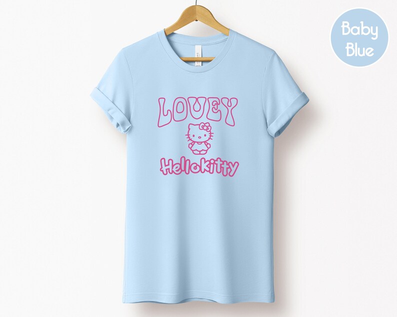 Op de afbeelding: Een lichtblauw T-shirt met opgerolde mouwen, hangend aan een houten hanger. Het shirt heeft de roze tekst "LOVEY" boven een Hello Kitty-afbeelding en de tekst "Hellokitty". Een lichtblauwe cirkel met "Baby Blue" staat rechtsboven.