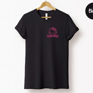 May include: Black t-shirt with a pink Hello Kitty graphic on the front. The t-shirt is hanging on a wooden hanger against a white background. The word "Black" is in a black circle in the upper right corner.