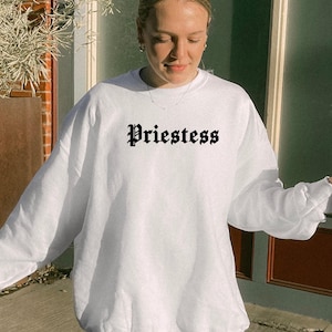 May include: White crew neck sweatshirt featuring the word "Priestess" in black gothic font. The sweatshirt has a relaxed fit and is likely made from a soft fabric. The design has a casual, comfortable style.