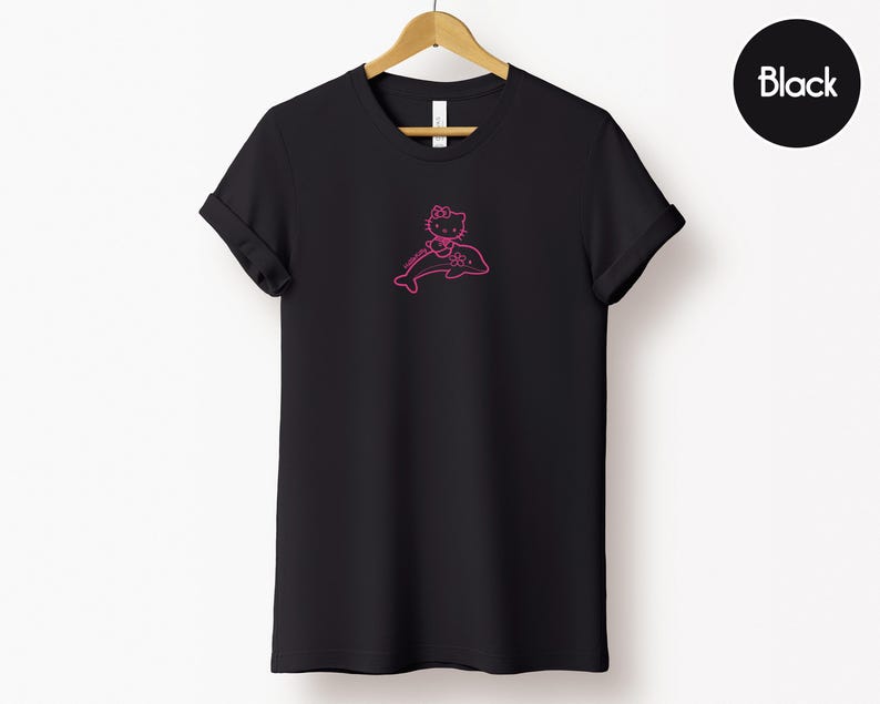 May include: Black t-shirt with a pink graphic of a cartoon cat riding a dolphin. The cat is wearing a bow and the dolphin is outlined in pink. The word "Black" is in a black circle in the upper right corner.